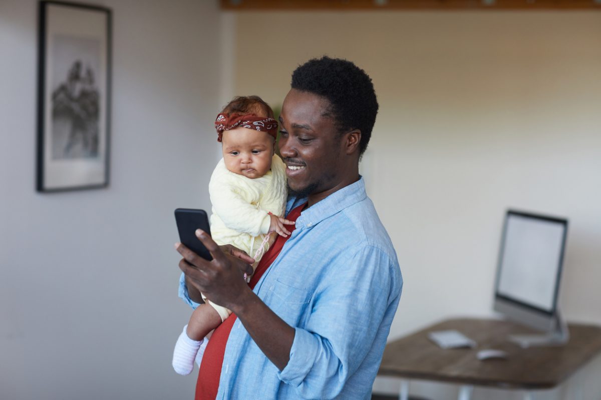 African smiling father holding baby girl on his hands and using his mobile phone at home