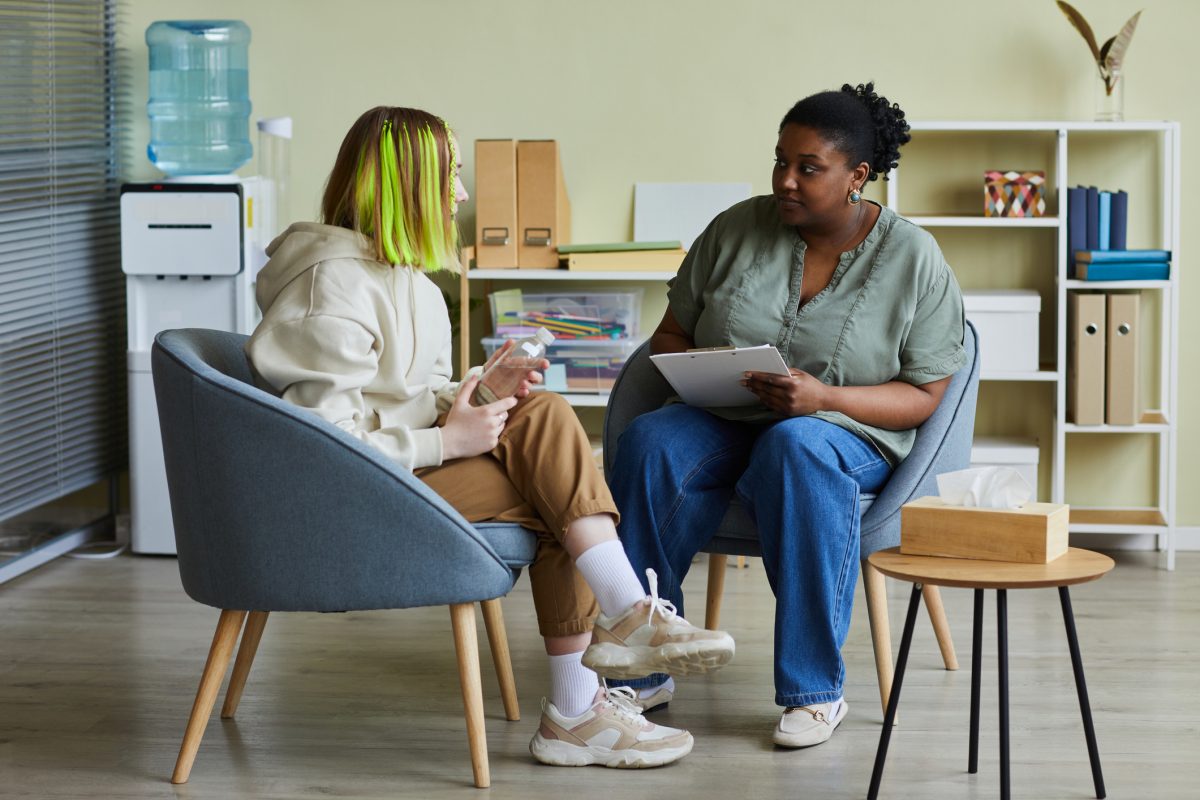 African female social worker talking to difficult teenage girl while they sitting on armchairs at office