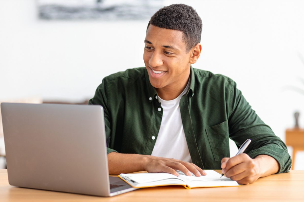 Smiling male student or freelancer studying remotely from home. African American male using laptop for online education, taking notes in a notebook. Online lesson, e-learning at home concept