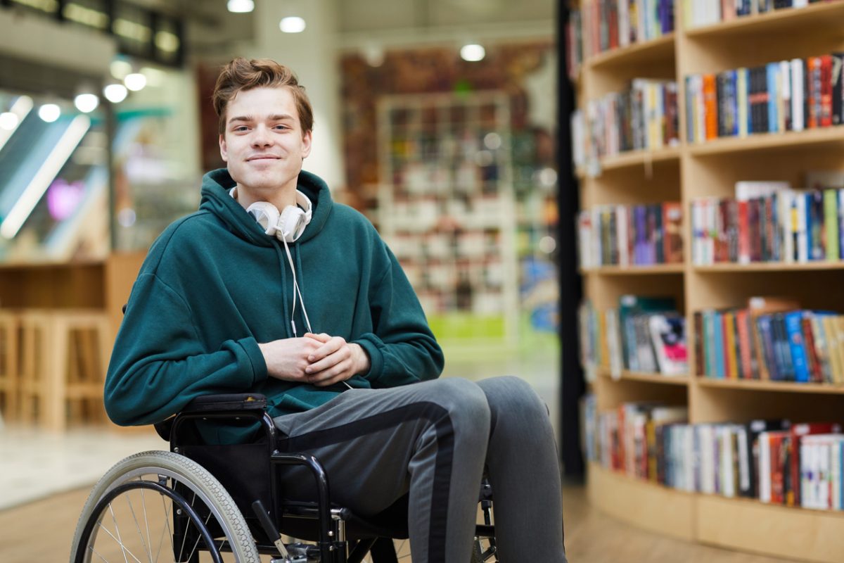 Content handsome young disabled student with headphones on neck siting in wheelchair and looking at camera in modern library or bookstore