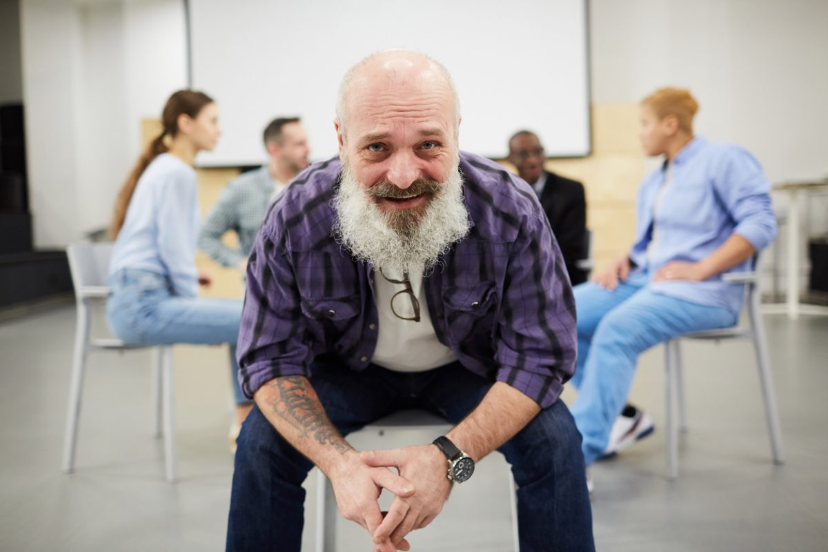 Portrait of bearded senior man smiling at camera while sitting in chair in front of therapy group, copy space