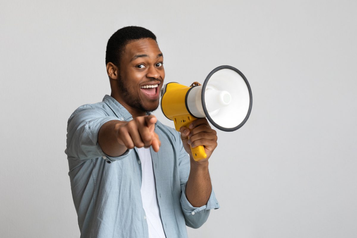 Positive african american guy shouting in megaphone and pointing at camera over grey background, copy space. Happy black man screaming with loudspeaker, cheering up, making advertisement