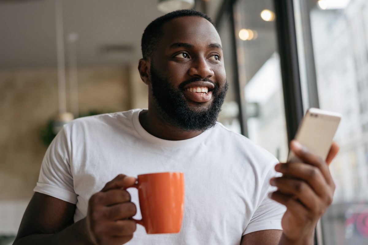 Portrait of handsome African American man using mobile phone, drinking coffee in cafe. Freelancer holding smartphone, working from home