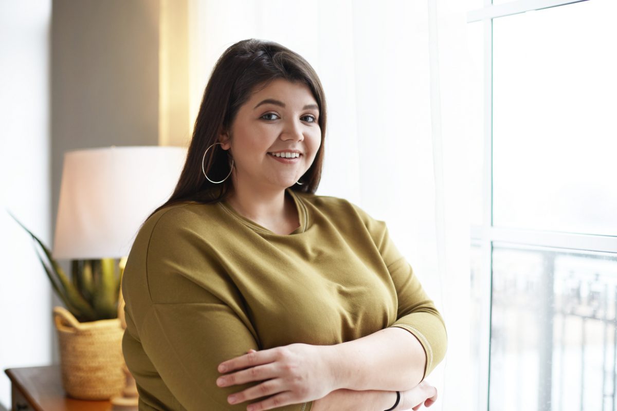 Portrait of attractive beautiful young female with chubby cheeks and curvy body posing at window at home, crossing arms on chest confidently and smiling at camera, enjoying leisure time indoors