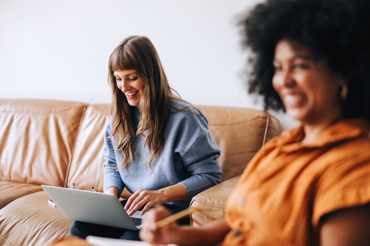 Happy young businesswoman using a laptop while sitting in an office lobby with her colleague. Cheerful young businesswomen working in a modern workplace.