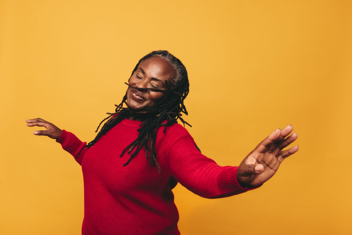 Joyful mature woman dancing and having fun while standing against a yellow background. Happy black woman with dreadlocks embracing her natural hair with pride.