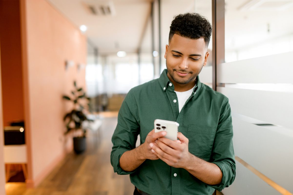 Handsome hispanic guy is using smartphone for messaging, texting, young indian man in smart casual green shirt using smartphone standing in office, typing message, chatting in the networks, scrolling